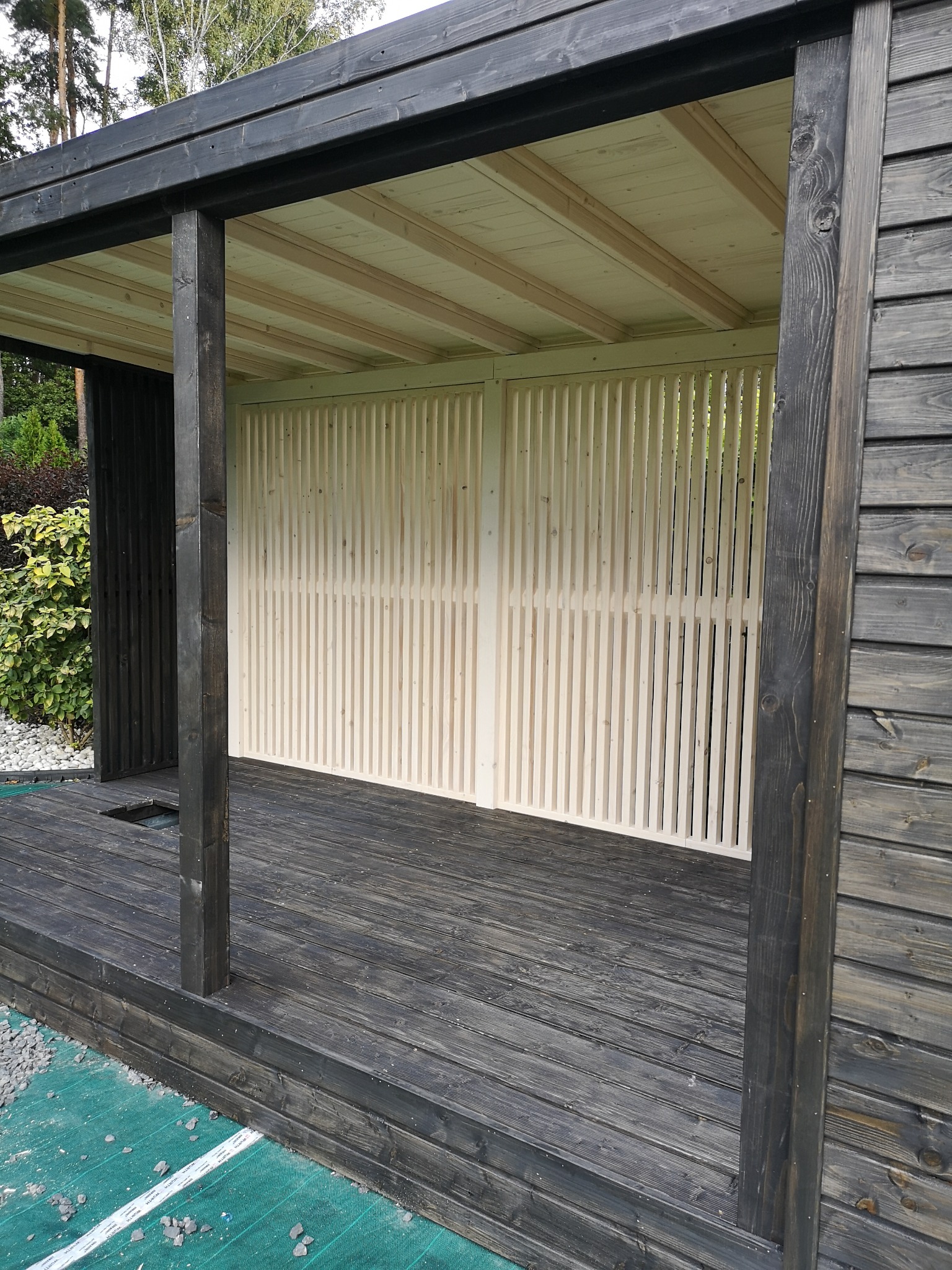 Nowoczesna konstrukcja z ciemnego drewna z jasnymi, pionowymi panelami i drewnianym podestem, widok z zewnątrz na tle zieleni.