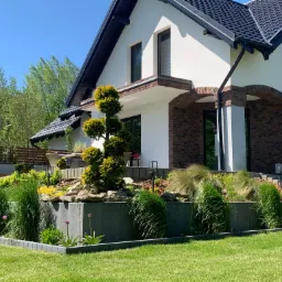 Nowoczesny dom z zadbanym ogrodem tarasowym, z kostki brukowej ułożone obrzeża rabat, ozdobne trawy i formowane drzewka iglaste.