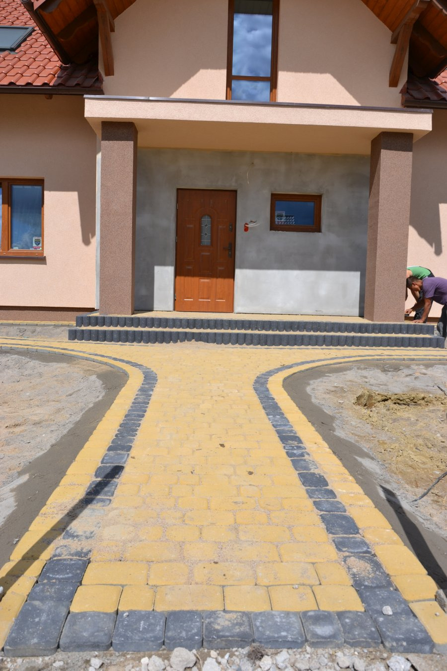 Żółto-szara kostka brukowa ułożona jako ścieżka do domu z brukarzem pracującym przy schodach wejściowych.