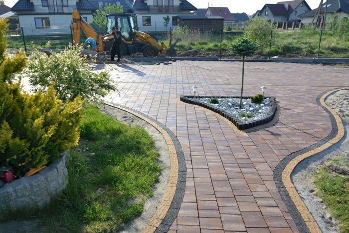 Ułożona kostka brukowa w odcieniach brązu tworząca ścieżkę i podjazd, z dekoracyjnym klombem w kształcie trójkąta wypełnionym białymi kamieniami i roślinami. W tle widoczny traktor wykonujący prace...