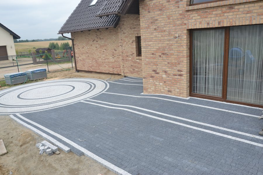 Ułożona szara kostka brukowa z białymi pasami i okrągłym wzorem przed ceglanym domem. Część kostki jeszcze nie ułożona, widoczne paczki kostki w tle.