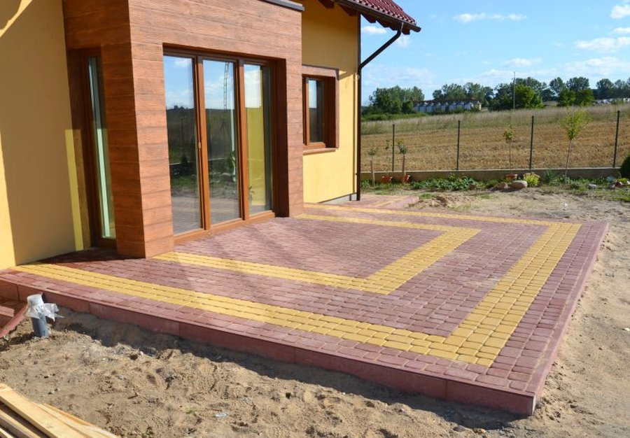 Wykończony taras z kostki brukowej w kolorach ceglastym i żółtym, przylegający do żółtego domu z brązowymi elementami wykończeniowymi, widok na ogrodzone pole w tle.