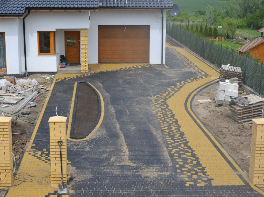 Częściowo ułożona, czarno-żółta kostka brukowa przed domem z brązową bramą garażową, widoczne przygotowane podłoże w kształcie łzy i ceglane słupki ogrodzeniowe z łopatą opartą o jeden z nich.