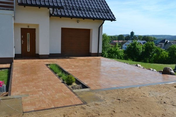 Nowo ułożona, brązowa kostka brukowa prowadząca do domu z garażem, z ozdobnym pasem zieleni pośrodku, widok na okoliczną zabudowę w tle.
