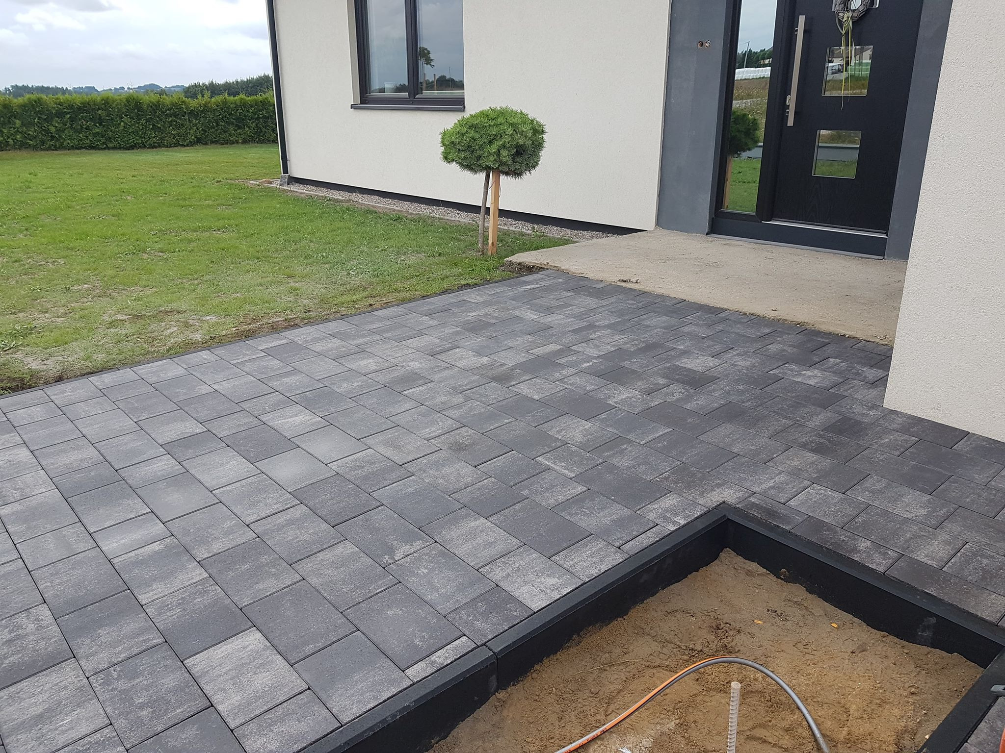 Ułożona kostka brukowa w odcieniach szarości przed nowoczesnym domem z czarnymi ramami okiennymi i drzwiami, widoczny fragment przygotowanego wykopu pod obrzeże z wystającą rurką.