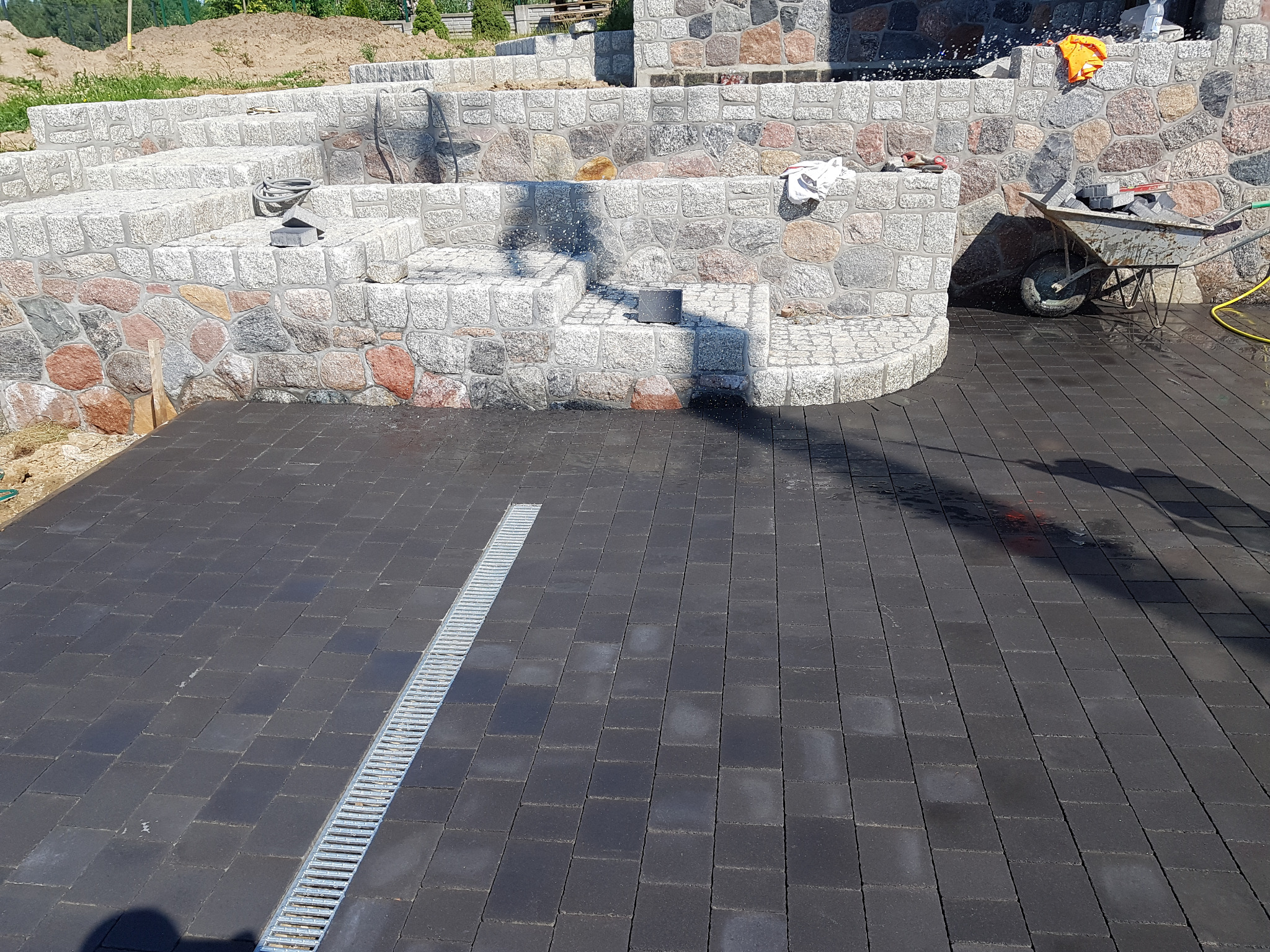 Ułożona, ciemnoszara kostka brukowa z odpływem liniowym na posesji, w tle kamienne schody i murki oporowe z elementami z jasnego granitu i ciemnych otoczaków, wózek budowlany z kostką i narzędziami.