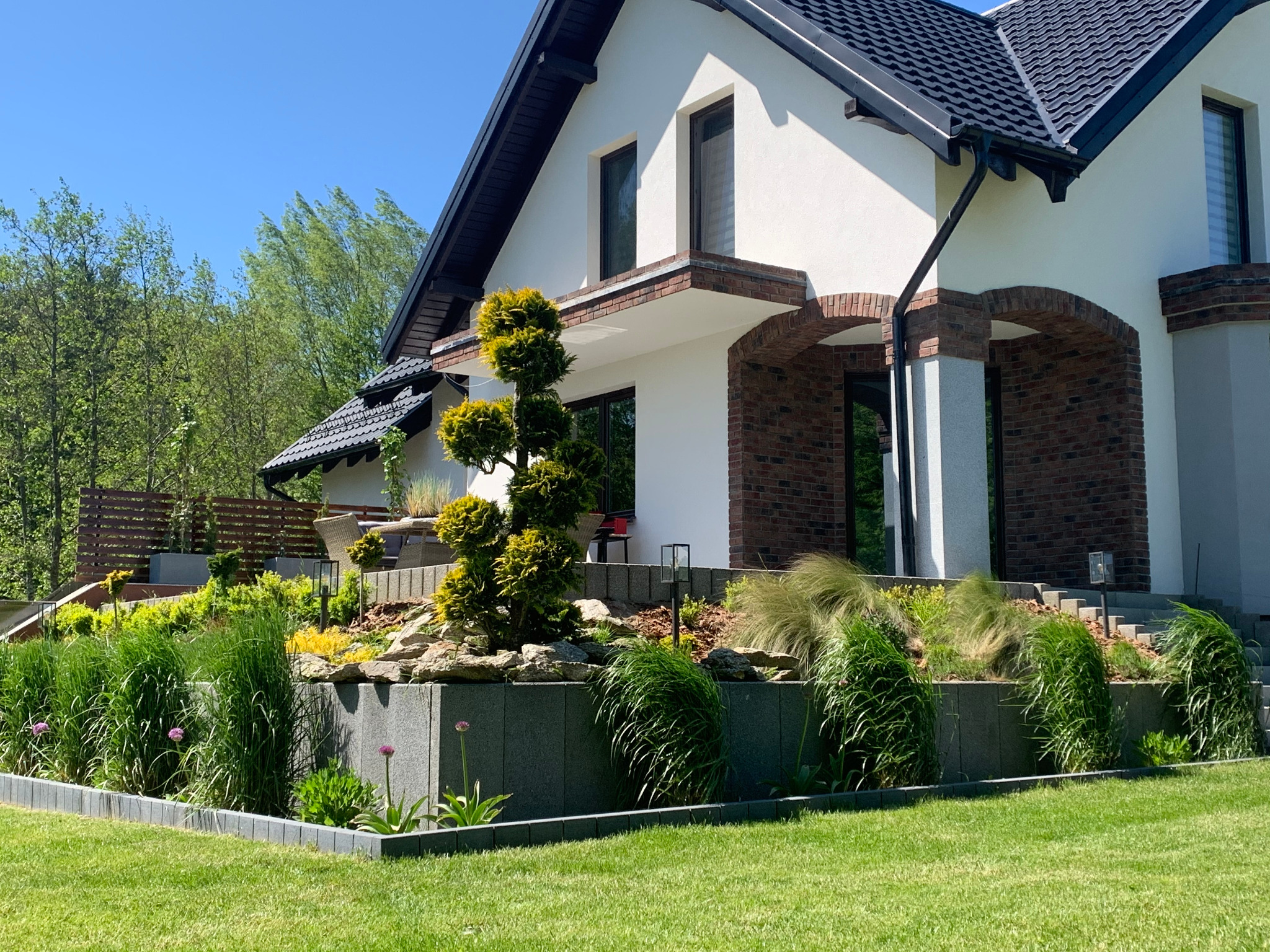 Nowoczesny dom z zadbanym ogrodem tarasowym, z kostki brukowej ułożone obrzeża rabat, ozdobne trawy i formowane drzewka iglaste.