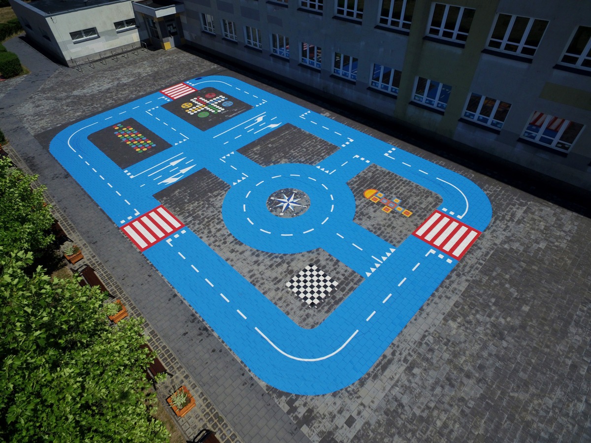 Nietypowy plac zabaw z namalowaną na kostce brukowej niebieską drogą z przejściami dla pieszych, rondem z kompasem, polami do gry w warcaby i chińczyka, całość sfotografowana z góry.
