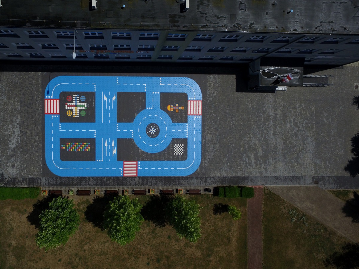 Widok z góry na niebieską strefę edukacyjną z namalowanymi drogami, przejściami dla pieszych i grami planszowymi na betonowym placu szkolnym, otoczoną zielenią i budynkiem.