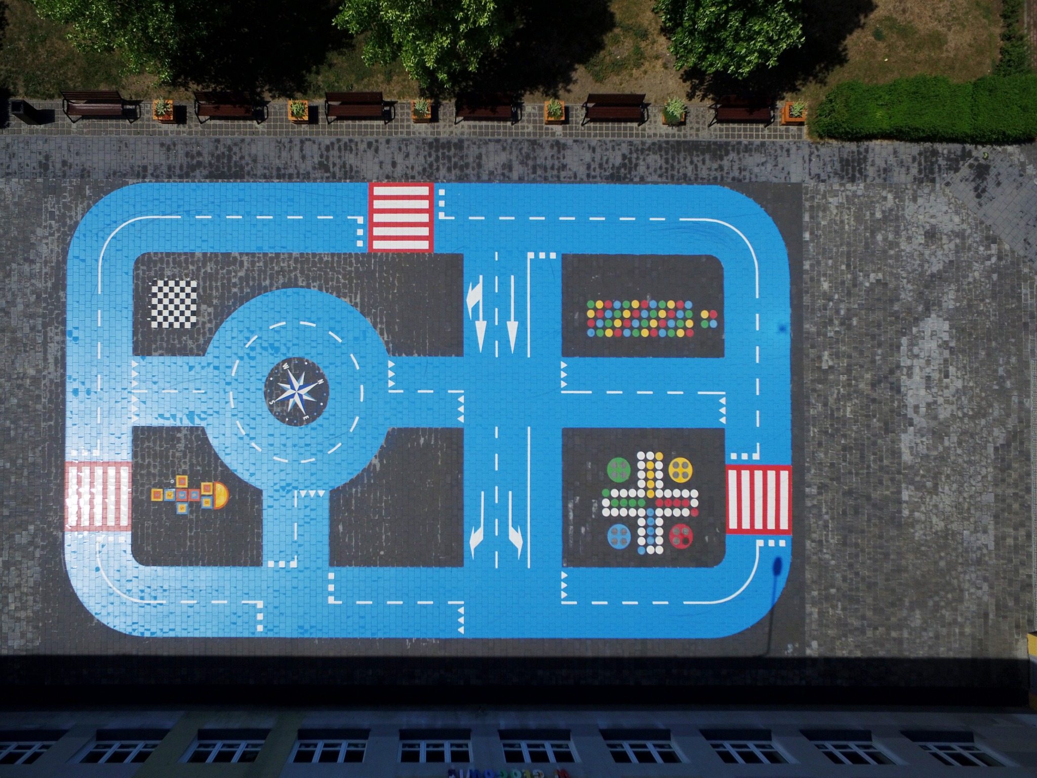 Widok z góry na niebieski plac zabaw z namalowanymi drogami, przejściami dla pieszych, rondem z kompasem i kolorowymi wzorami na szarym bruku, otoczony zielenią i ławkami.