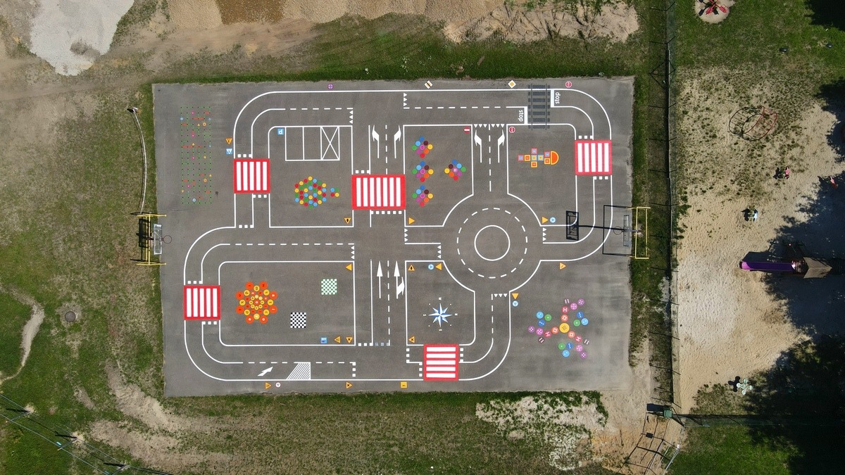 Widok z góry na plac zabaw z namalowaną na asfalcie imitacją ulicy z przejściami dla pieszych, rondem, miejscami parkingowymi i różnokolorowymi elementami dekoracyjnymi.