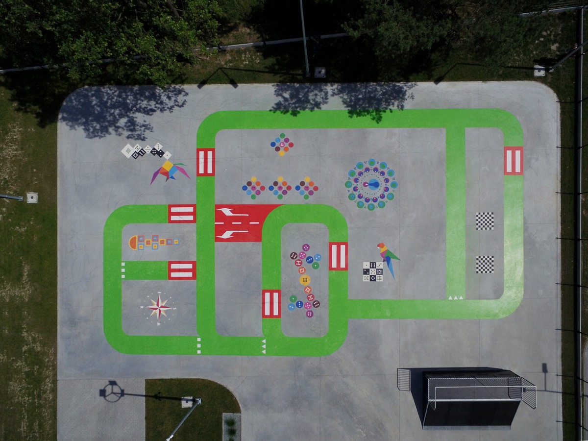 Widok z góry na plac z betonową nawierzchnią, na której namalowano zielone ścieżki i kolorowe wzory, w tym kostki do gry, mandalę, kompas i szachownicę.