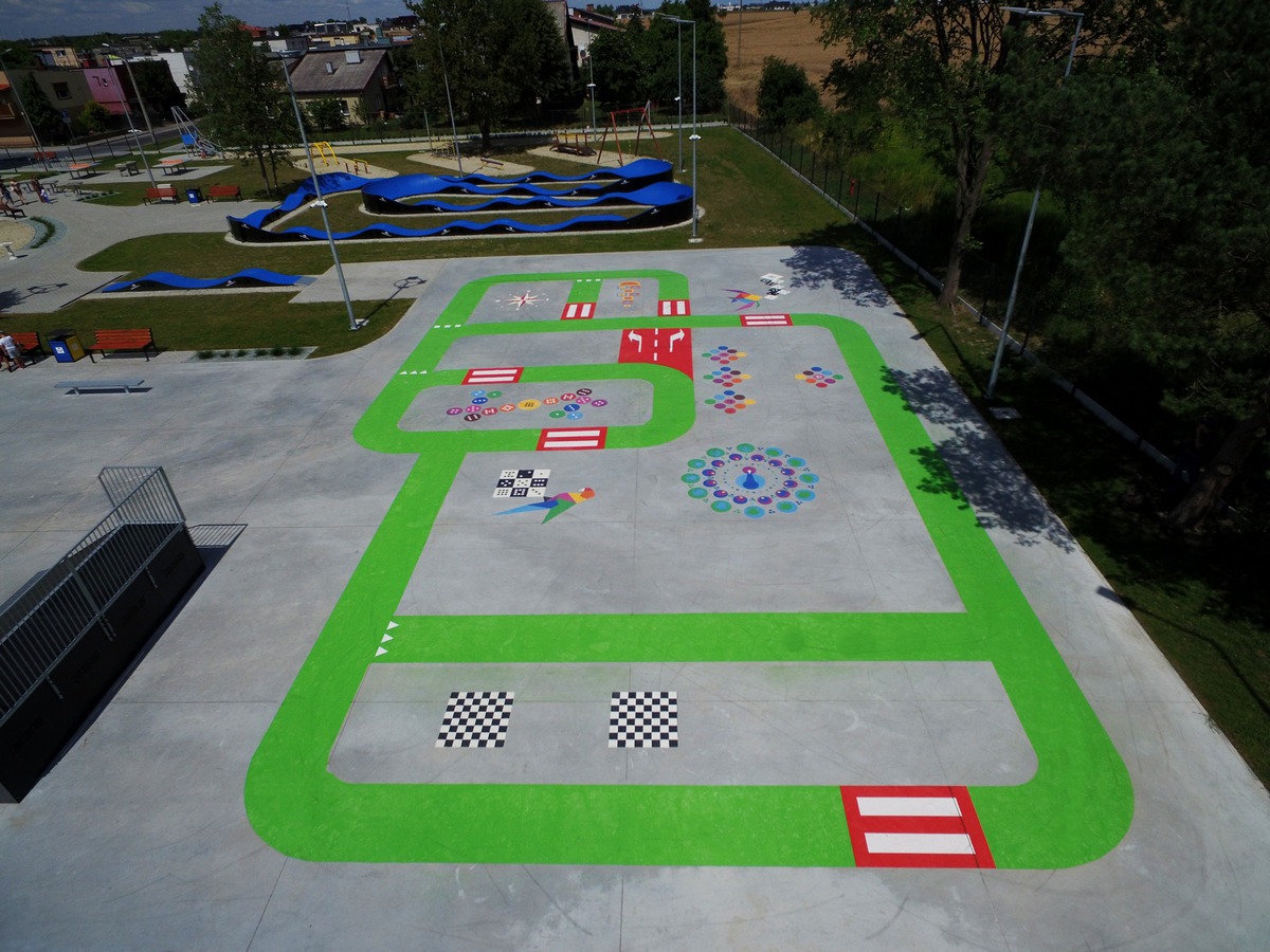 Widok z góry na betonowy plac zabaw z namalowaną zieloną trasą z kolorowymi elementami edukacyjnymi, w tle niebieski tor pumptrack i drzewa.