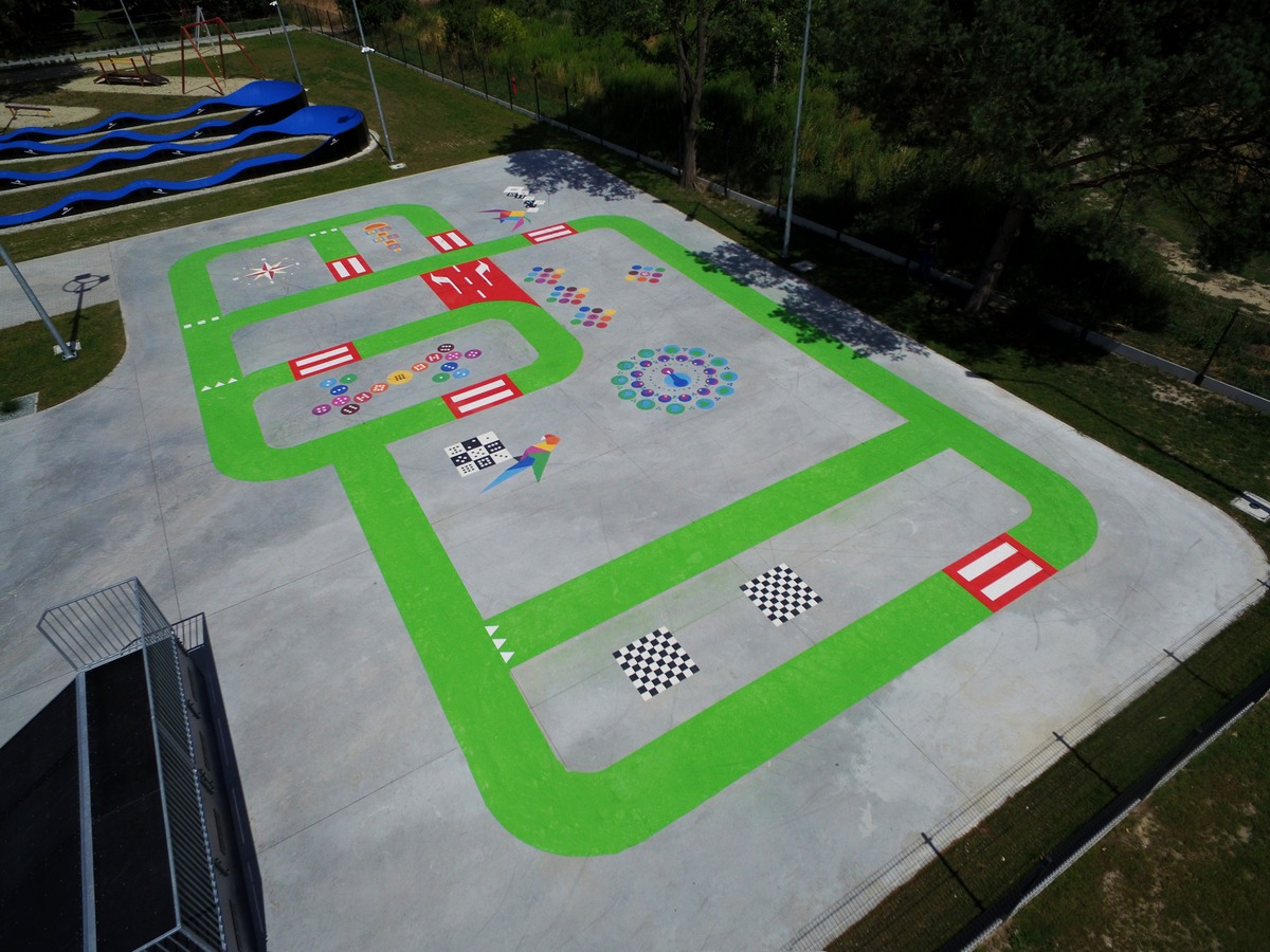 Widok z góry na betonowy plac zabaw z zielonym torem, kolorowymi malowidłami, elementami edukacyjnymi i huśtawkami w tle, oświetlony słonecznym światłem.
