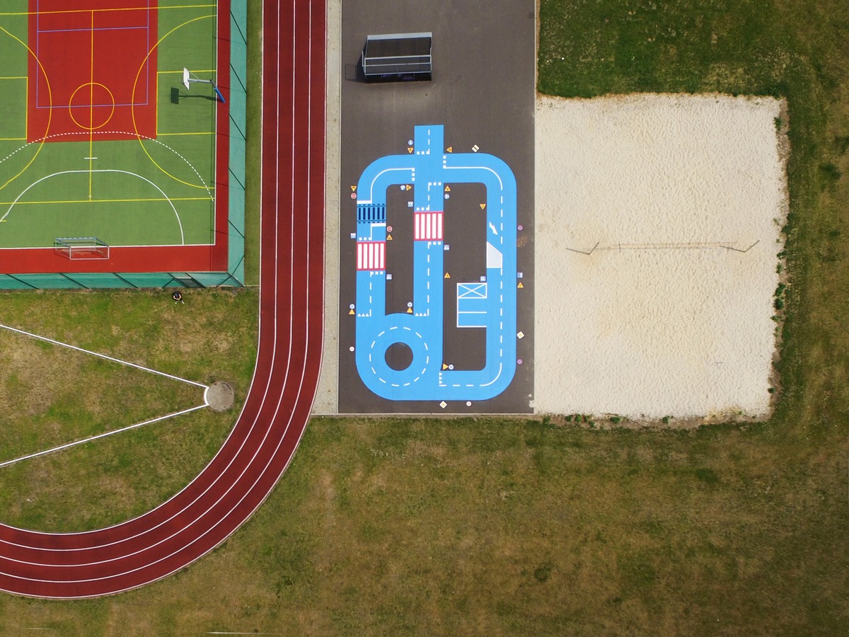 Widok z góry na plac zabaw z namalowaną niebieską trasą samochodową, obok bieżnia lekkoatletyczna i boisko sportowe.