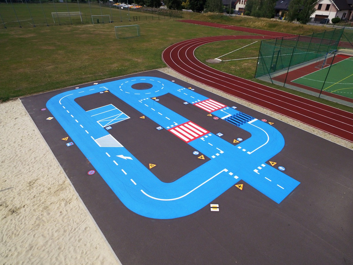 Nawierzchnia placu zabaw z namalowanym torem edukacyjnym dla dzieci, imitującym drogę z przejściami dla pieszych i znakami drogowymi, widok z góry.