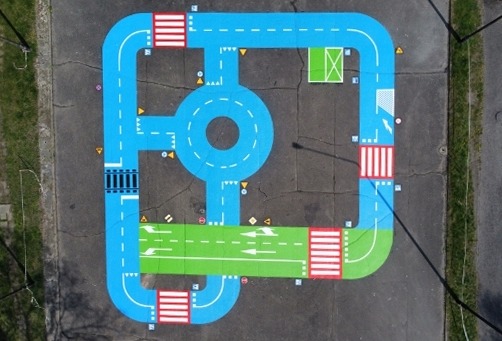 Nawierzchnia placu zabaw z namalowanym torem edukacyjnym dla dzieci, imitującym ruch drogowy z rondem, przejściami dla pieszych, torami kolejowymi i znakami drogowymi, widok z góry.