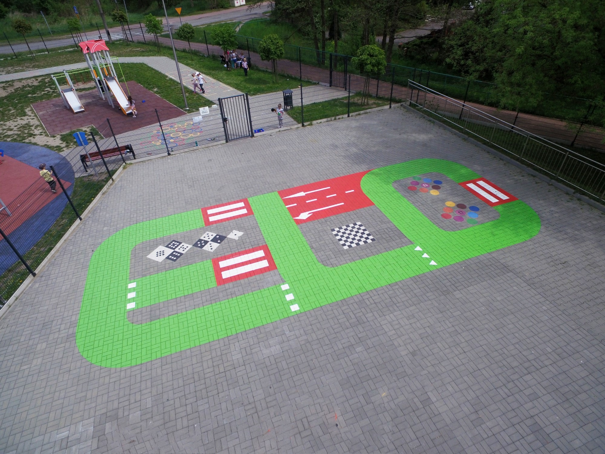 Z góry widoczny plac zabaw z namalowaną na szarym bruku trasą z zielonymi obrzeżami, czerwonymi strefami i białymi znakami, obok huśtawki i zjeżdżalnie, w tle grupa dzieci.
