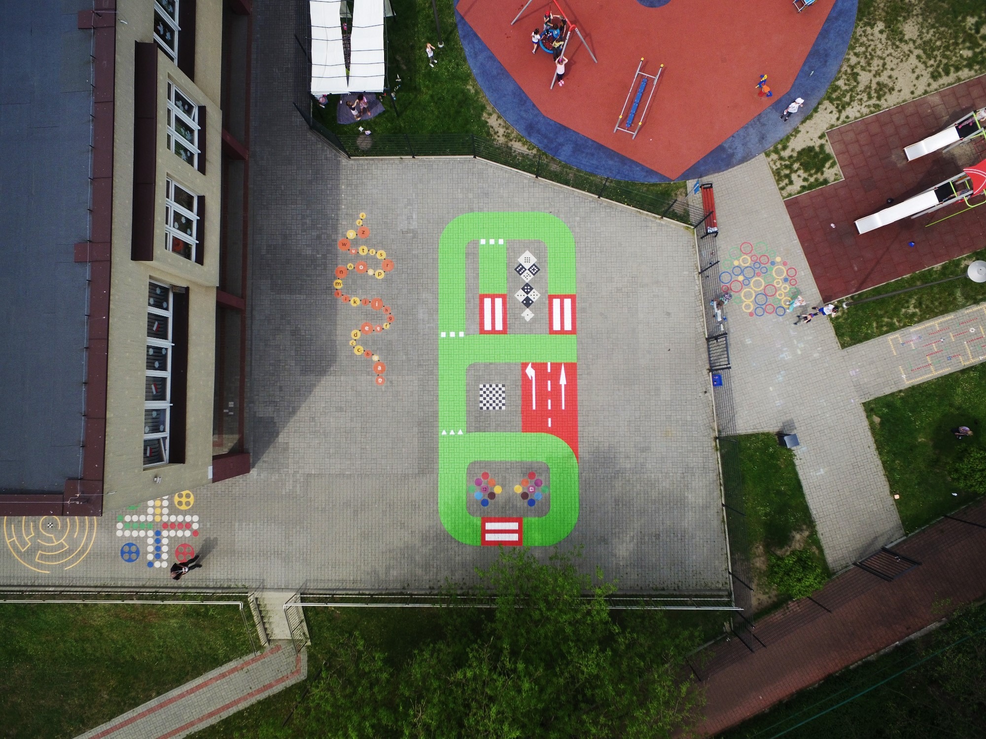 Widok z góry na plac zabaw szkolny z kolorowymi malowidłami na nawierzchni, w tym gra planszowa, ścieżka edukacyjna i tor przeszkód.