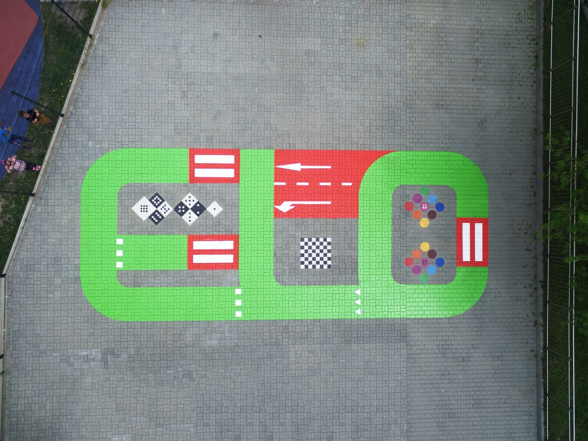 Widok z góry na pomalowaną nawierzchnię placu zabaw z elementami imitującymi drogę, kostki do gry i kolorowe koła, otoczoną szarą kostką brukową.