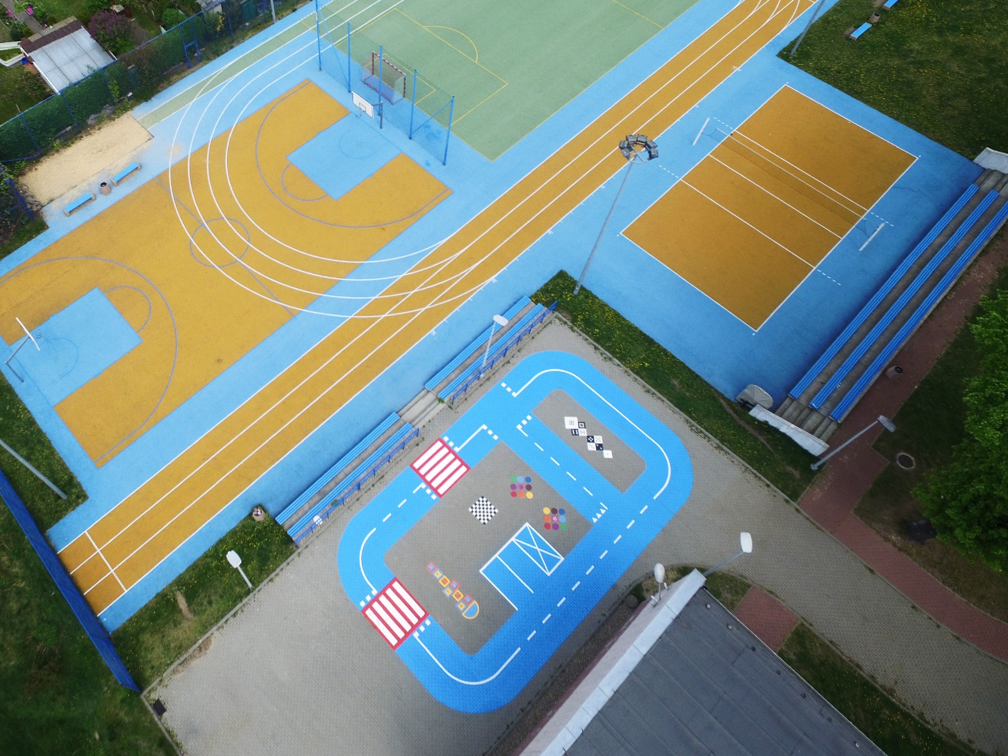 Widok z góry na kolorowy plac zabaw z bieżnią, boiskiem i edukacyjną strefą z namalowanymi grami drogowymi na szarym podłożu.