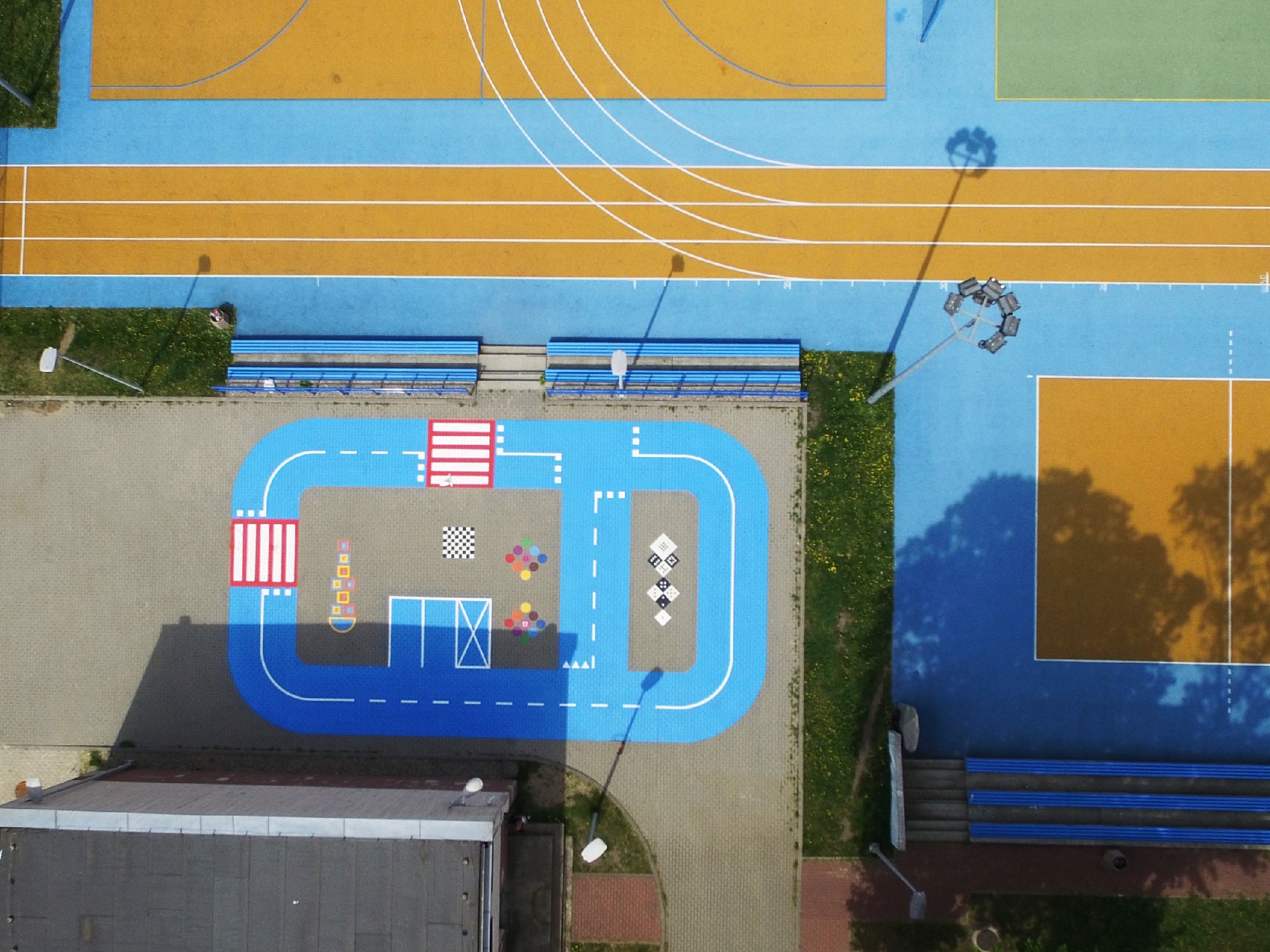 Widok z góry na kolorowy plac zabaw z elementami edukacyjnymi, w tym tor z przejściami dla pieszych, szachownice i inne gry, zintegrowany z boiskiem sportowym i trybunami.