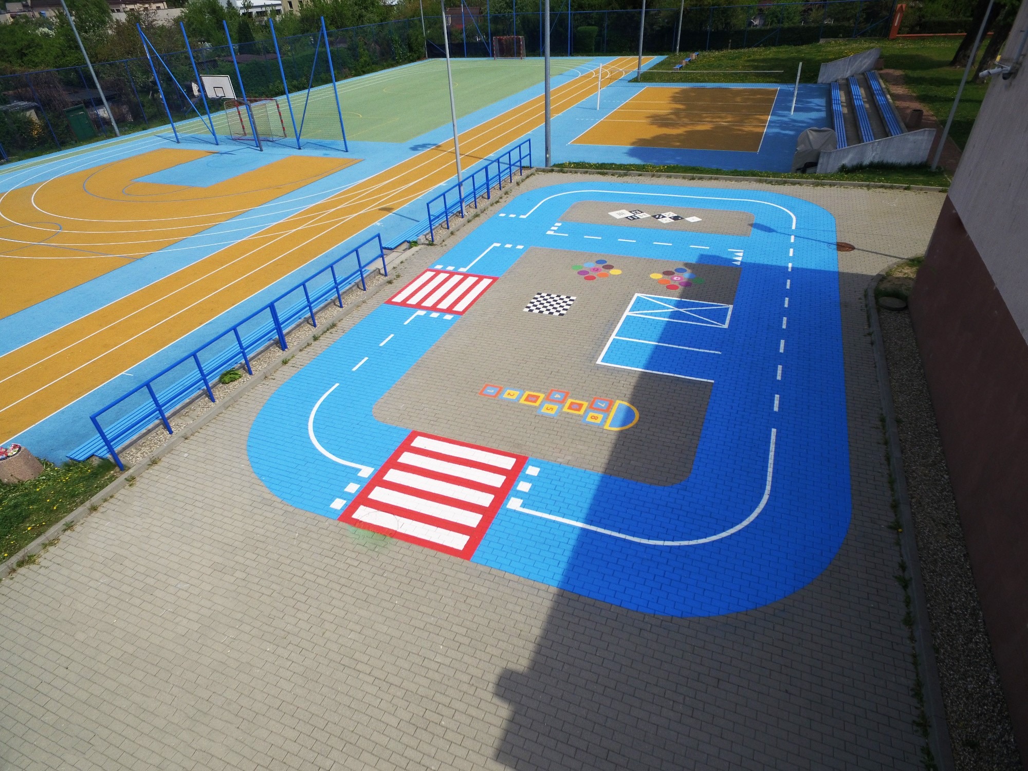 Kolorowa nawierzchnia placu zabaw z namalowaną trasą dla dzieci, zebrami, polami do gier i bieżnią w tle.