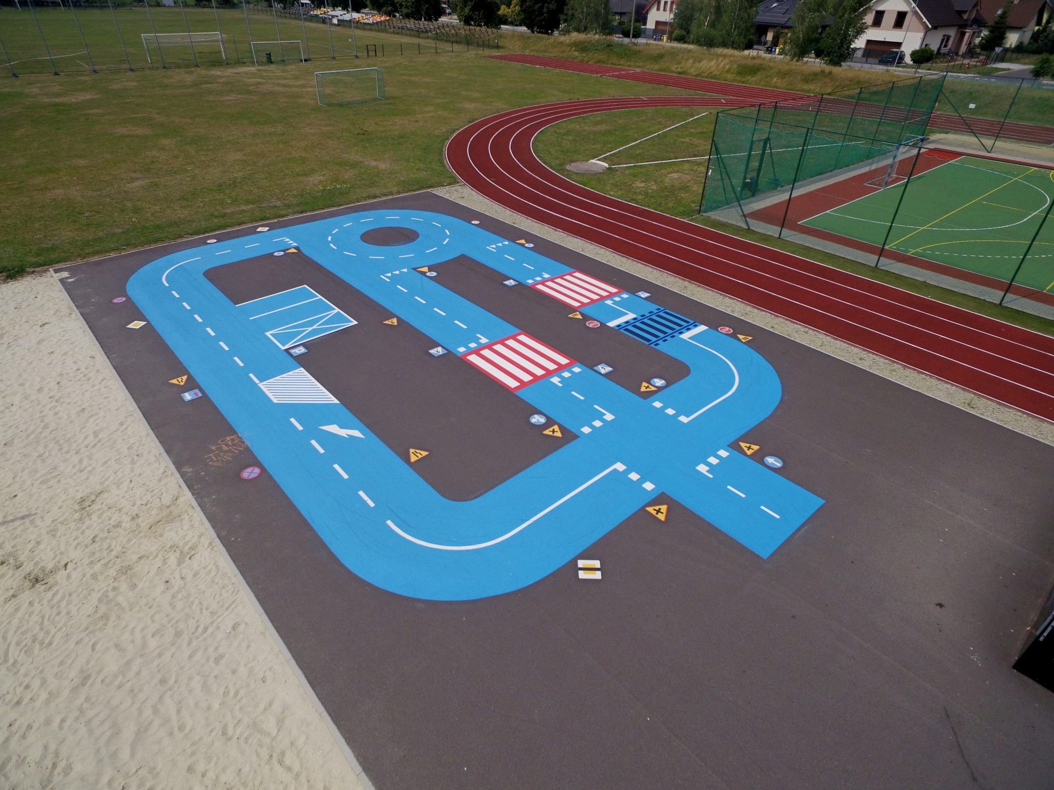 Nawierzchnia z namalowanym torem samochodowym, imitującym ulicę z przejściami dla pieszych i znakami drogowymi, obok bieżnia i boisko sportowe.