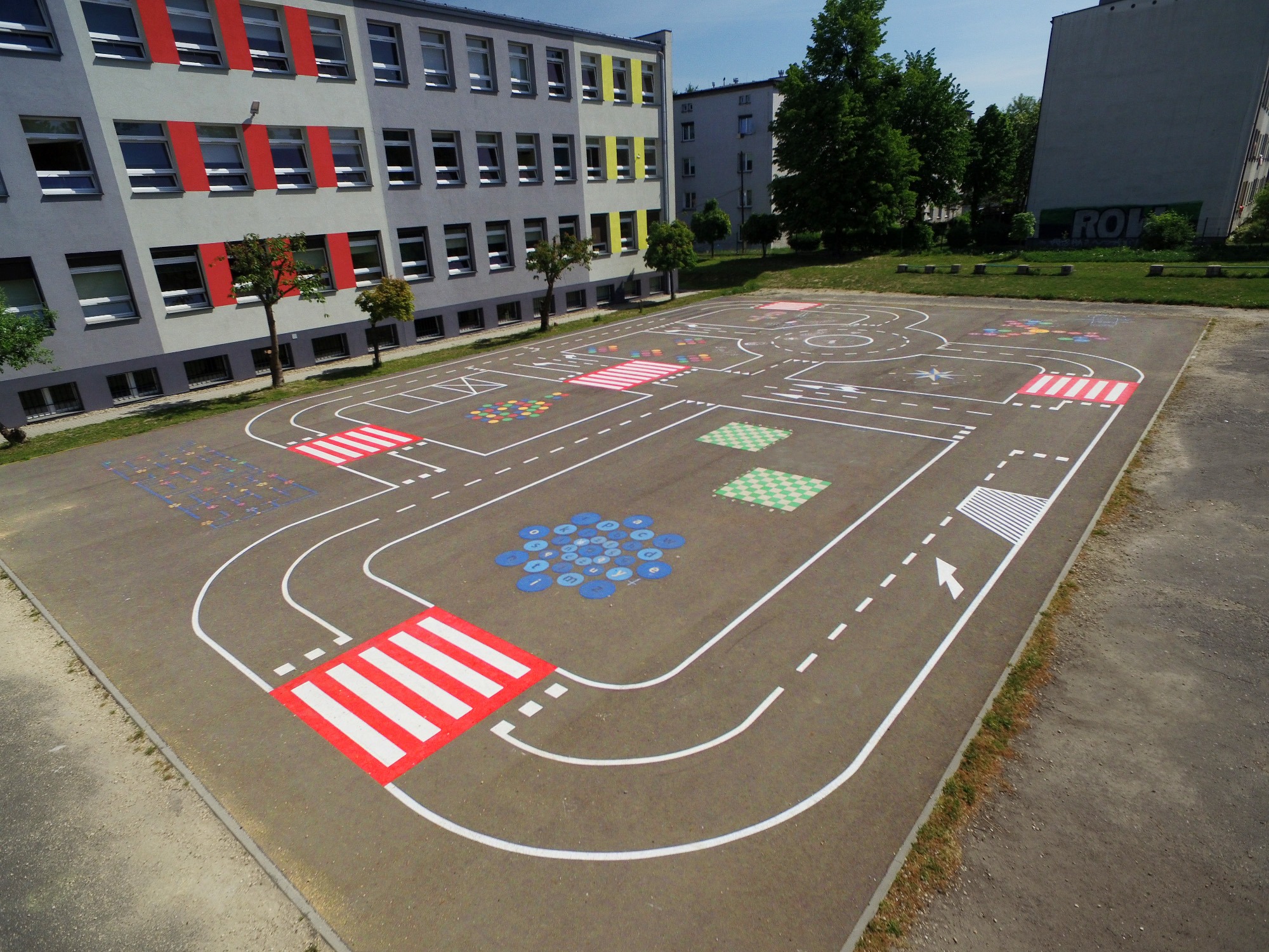 Oznakowanie poziome na asfalcie przedstawiające miasteczko ruchu drogowego z przejściami dla pieszych, drogami i elementami edukacyjnymi, na tle budynku szkoły z czerwonymi i żółtymi akcentami.