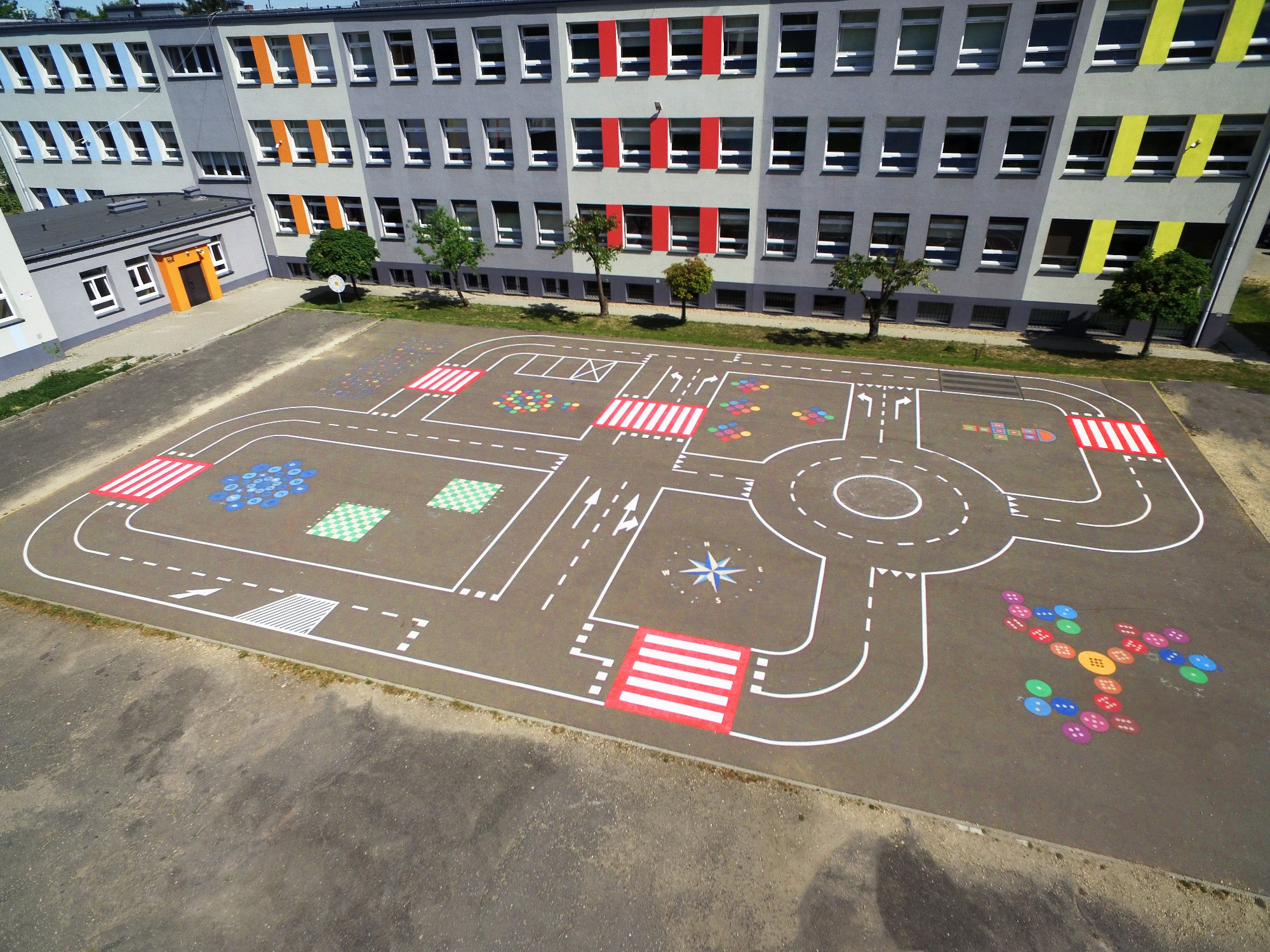 Malowany na asfalcie tor edukacyjny z przejściami dla pieszych, rondem, strzałkami kierunkowymi i kolorowymi polami, na tle budynku szkoły.