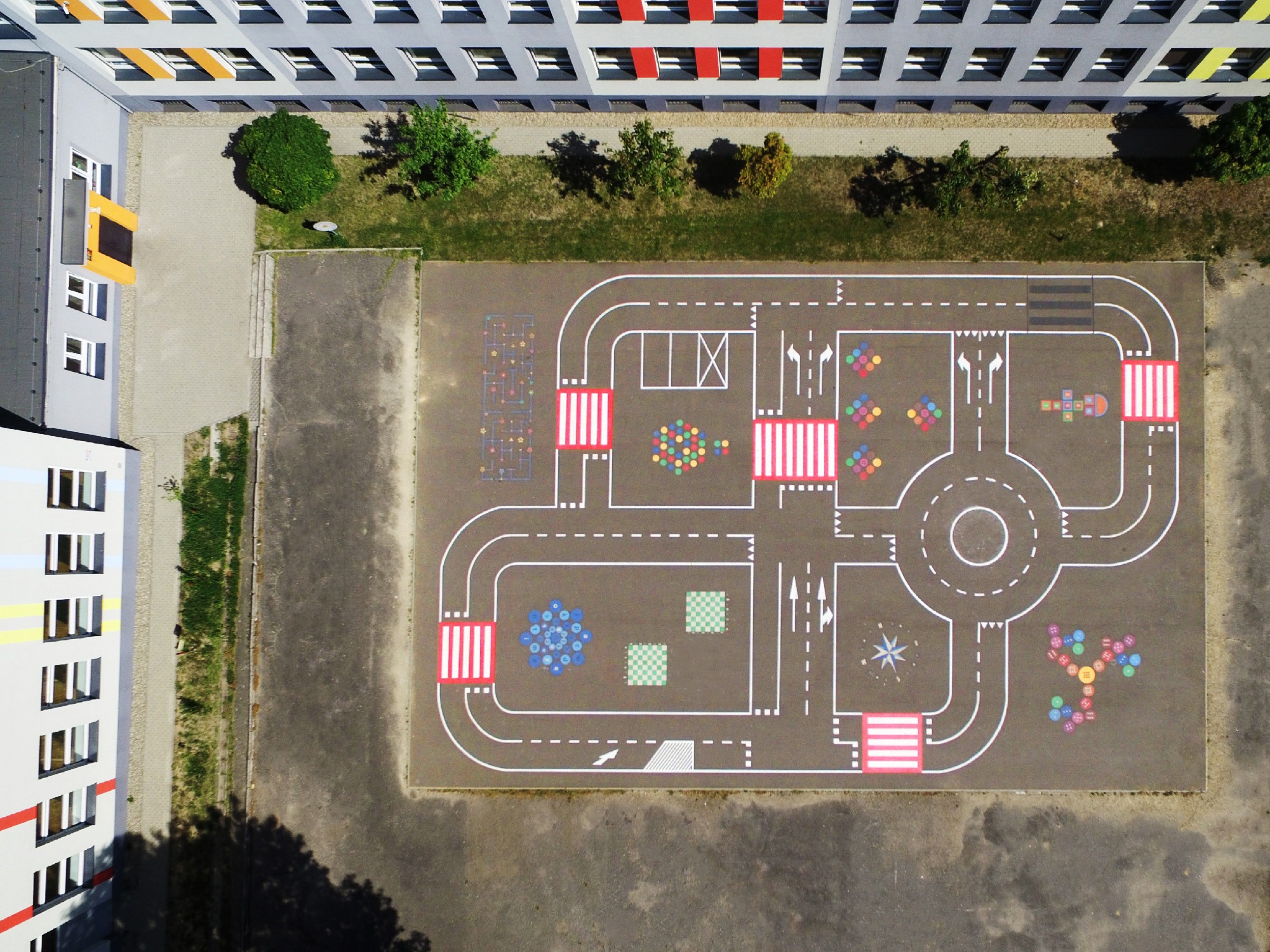 Widok z góry na plac zabaw z namalowanymi na asfalcie elementami miasteczka ruchu drogowego, w tle budynek szkoły.