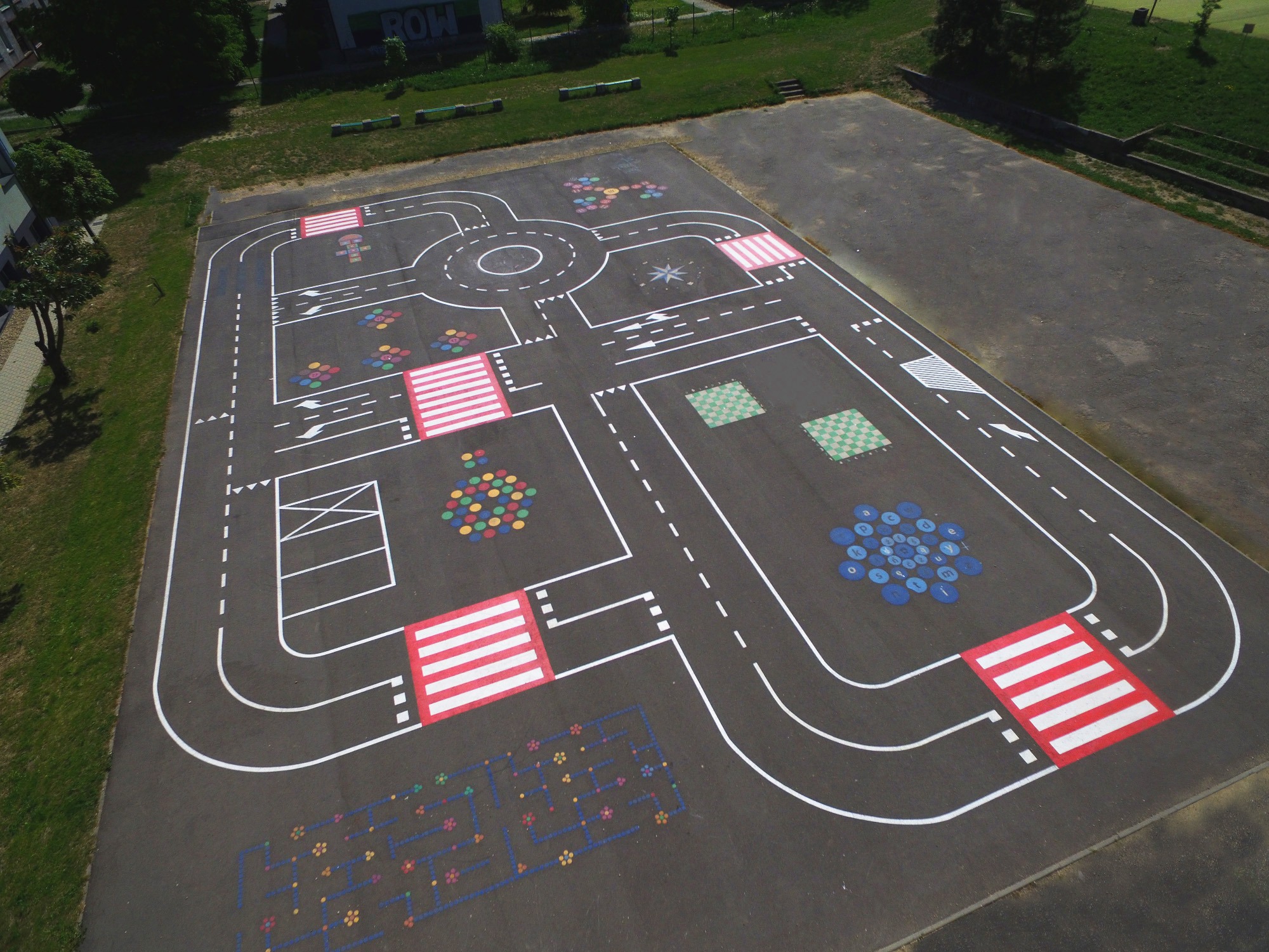 Malowany na asfalcie miasteczko ruchu drogowego z przejściami dla pieszych, rondem, parkingiem i kolorowymi elementami edukacyjnymi, widok z góry.