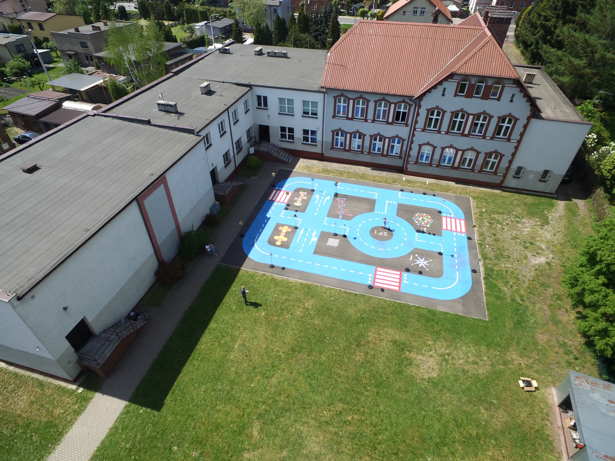 Widok z góry na szkolne podwórko z namalowanym niebieskim torem rowerowym i przejściami dla pieszych, otoczone budynkami szkolnymi i zielenią.