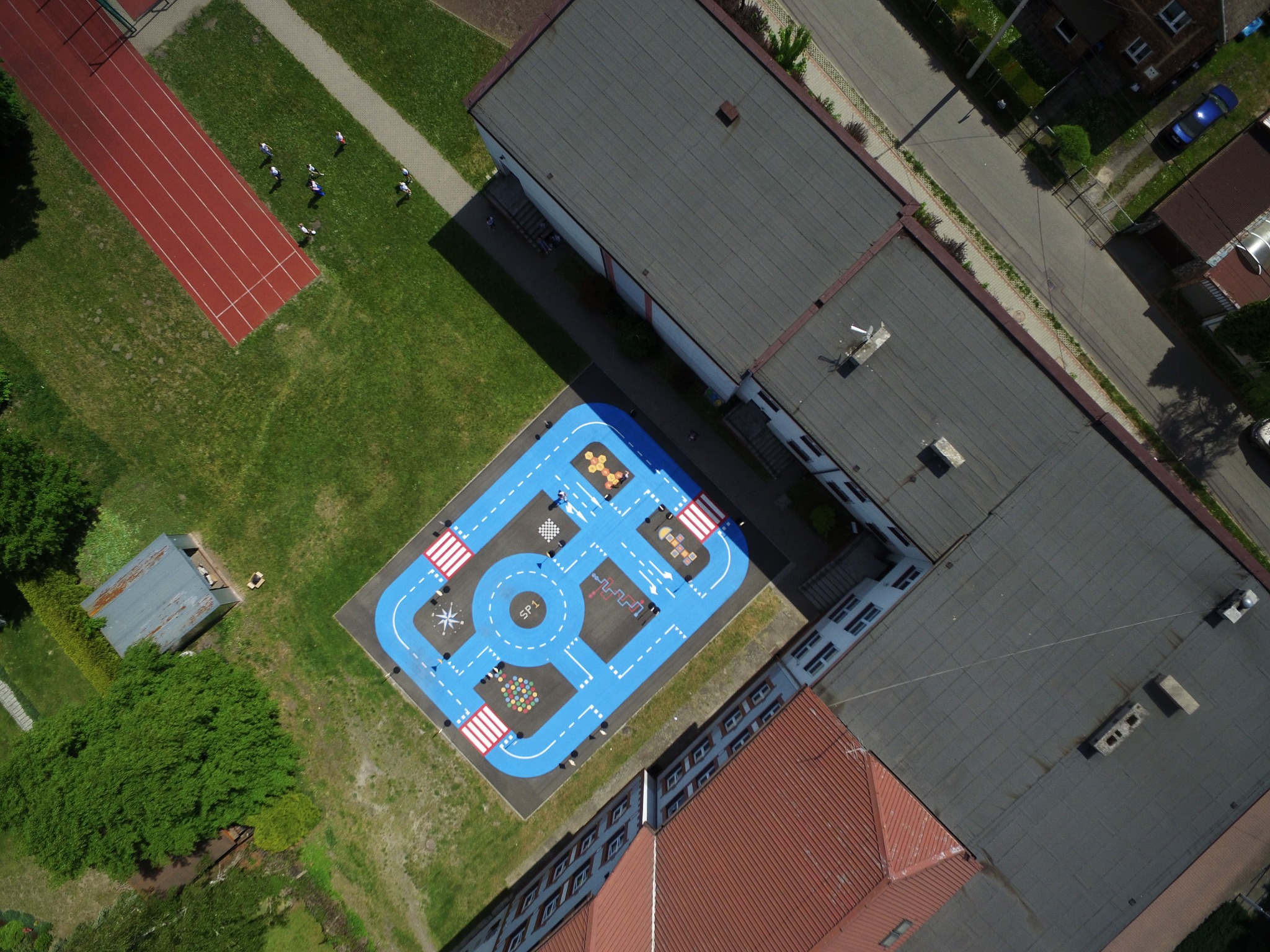 Widok z góry na niebieski plac zabaw w kształcie toru samochodowego z namalowanymi znakami drogowymi i elementami edukacyjnymi, przylegający do budynku szkoły z czerwonym dachem, obok bieżnia...