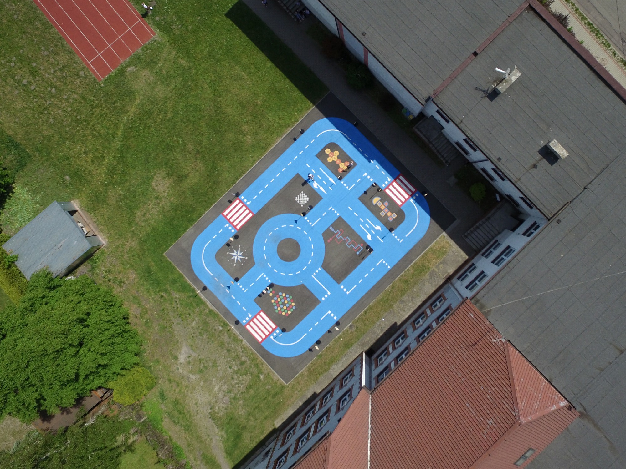 Widok z góry na plac zabaw z namalowaną niebieską ścieżką imitującą drogę, z przejściami dla pieszych i różnymi elementami edukacyjnymi, otoczony zielenią i budynkami.