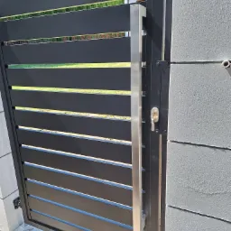 Nowoczesna brama wjazdowa z poziomymi, czarnymi panelami, stalowym uchwytem i zamkiem, zamontowana na betonowym słupku.