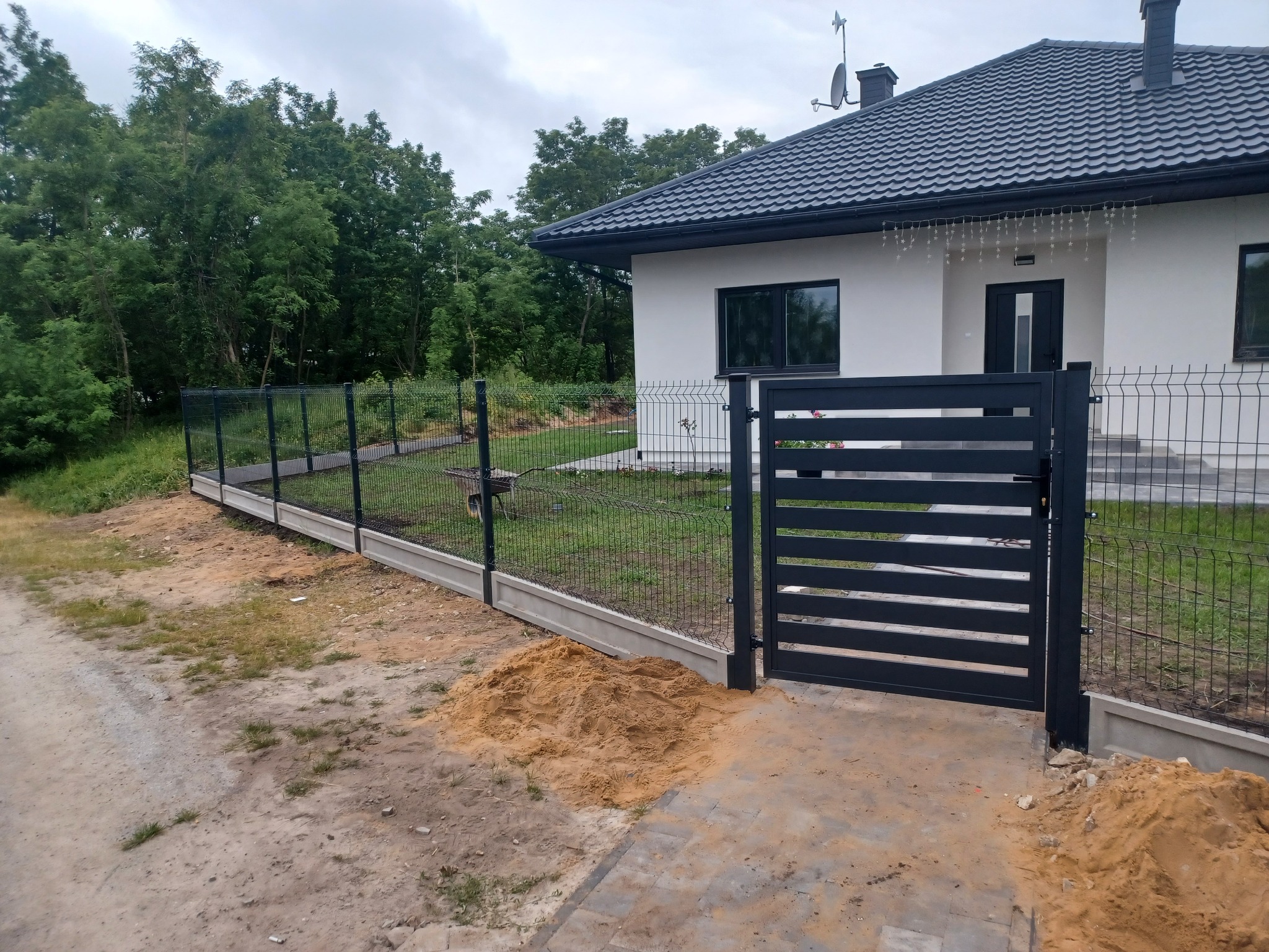 Nowoczesna posesja z czarną bramą wjazdową i ogrodzeniem panelowym na betonowej podmurówce, widoczny stos piasku przed bramą, w tle biały dom z szarą dachówką i zielone drzewa.