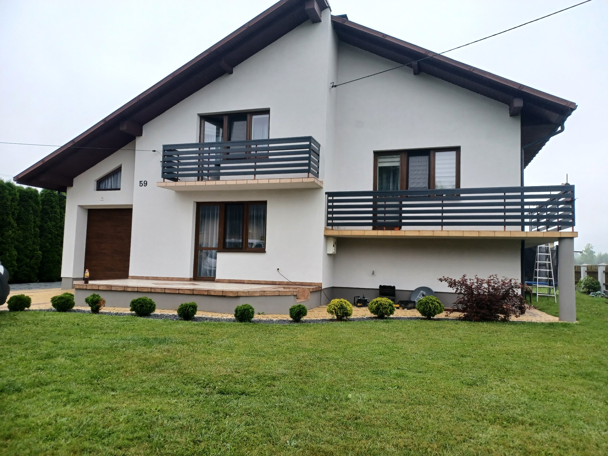 Nowoczesny dom jednorodzinny z białymi ścianami i ciemnobrązowymi ramami okiennymi oraz balkonami z poziomymi, czarnymi barierkami, widok z zadbanego trawnika z ozdobnymi krzewami.