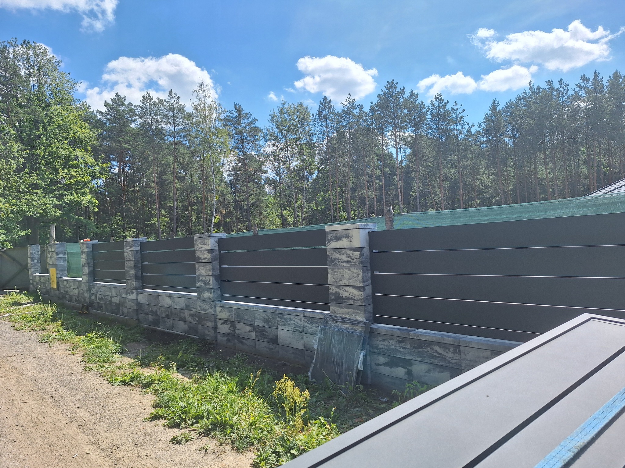 Nowoczesne ogrodzenie z poziomych paneli w kolorze grafitowym, osadzone na kamiennej podmurówce i słupkach, z widoczną siatką maskującą w tle i fragmentem otwartej bramy wjazdowej z boku.