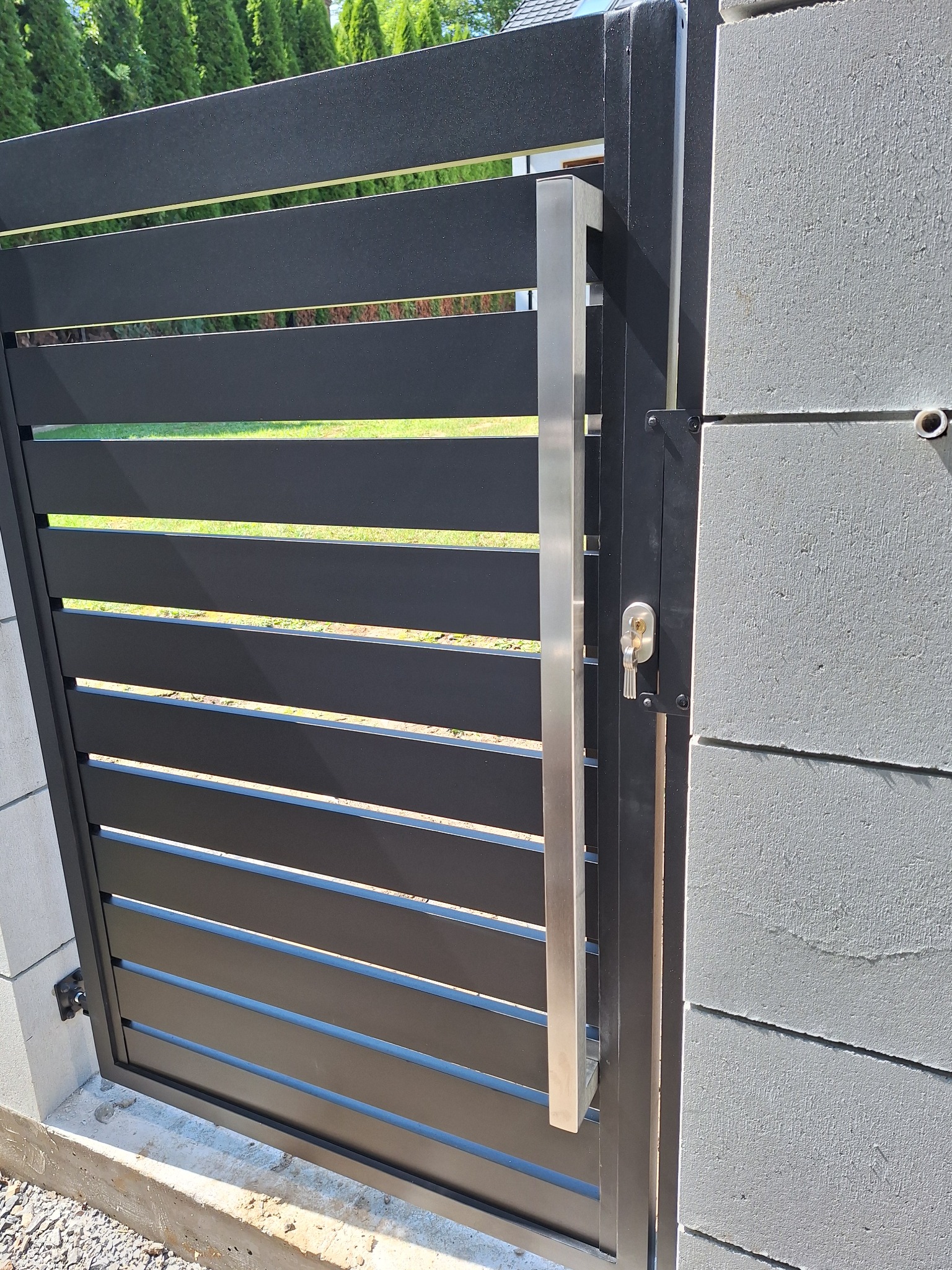 Nowoczesna brama wjazdowa z poziomymi, czarnymi panelami, stalowym uchwytem i zamkiem, zamontowana na betonowym słupku.