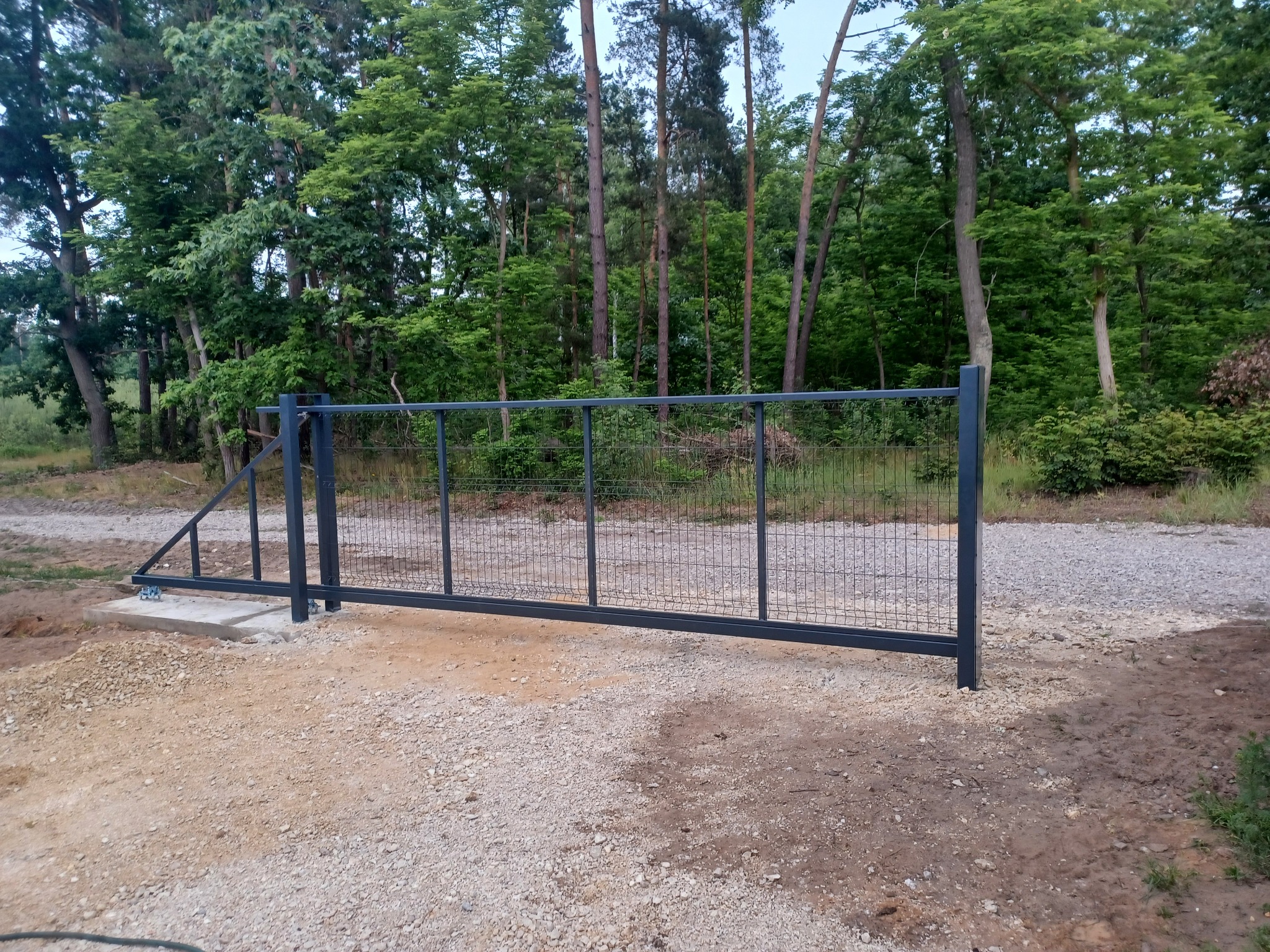 Metalowa brama wjazdowa przesuwna, szara, z siatkowym wypełnieniem, zamontowana na betonowej podstawie, w tle las.