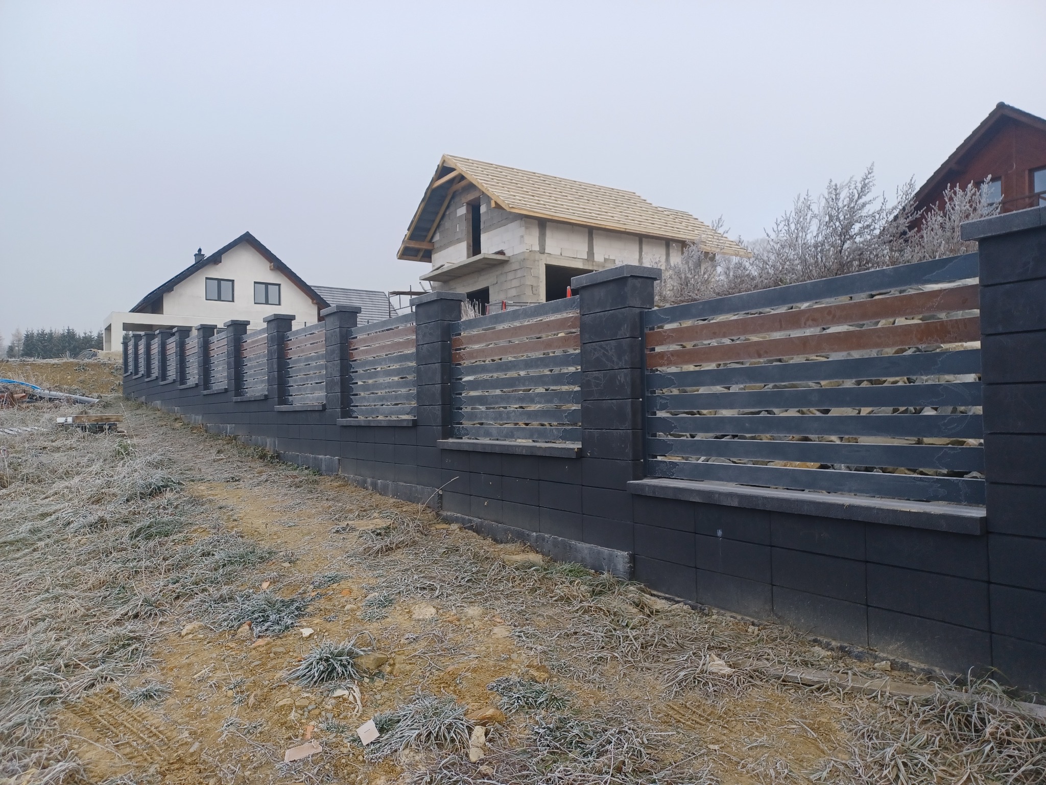 Długi, nowoczesny płot z szarych bloczków betonowych i poziomych, brązowo-szarych paneli, widoczny na tle budynków w różnym stadium budowy, w zimowej scenerii z oszronioną trawą.