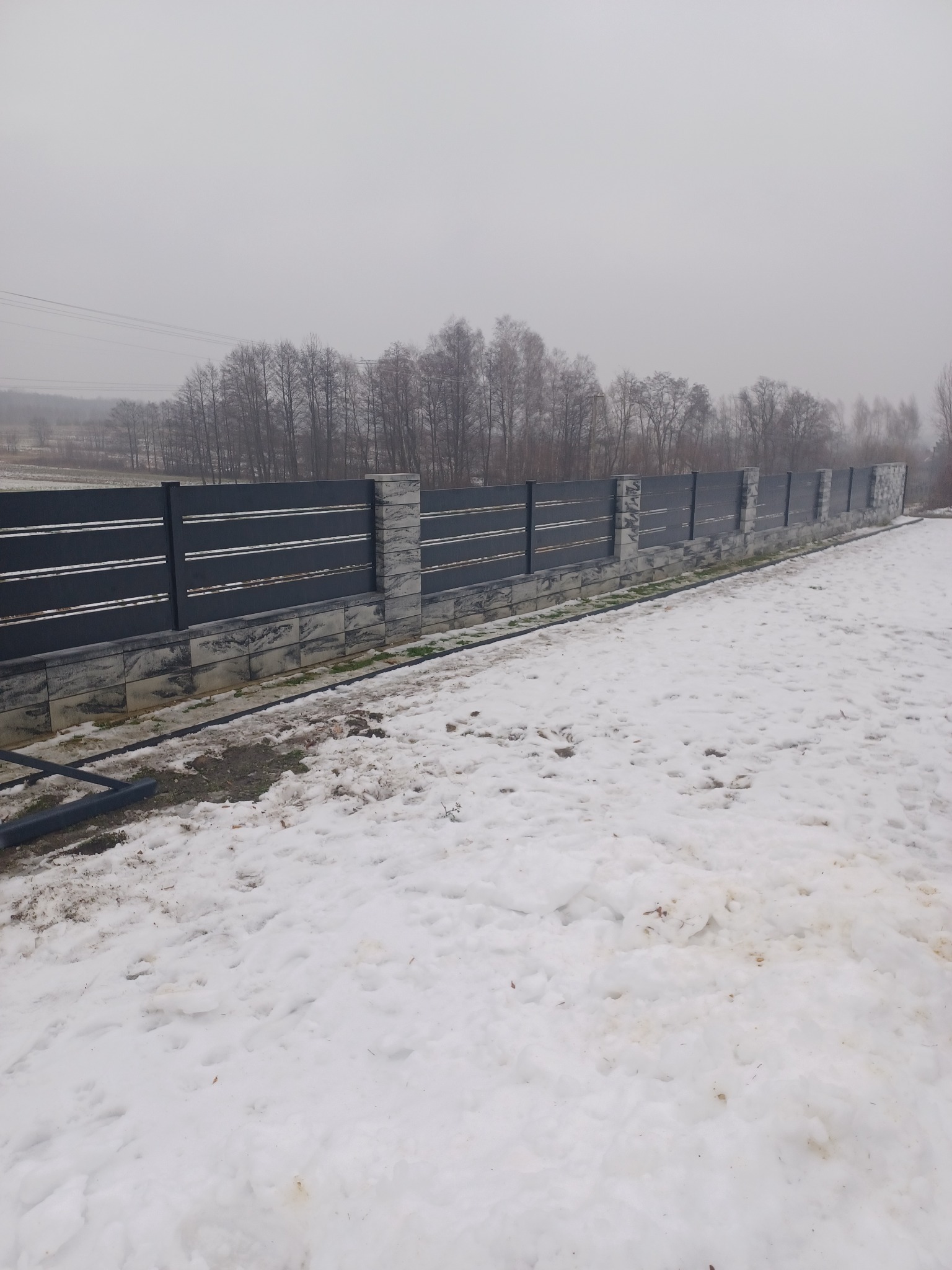 Nowoczesne, poziome ogrodzenie z szarych paneli i kamiennych słupków w zimowym krajobrazie, częściowo pokryte śniegiem.
