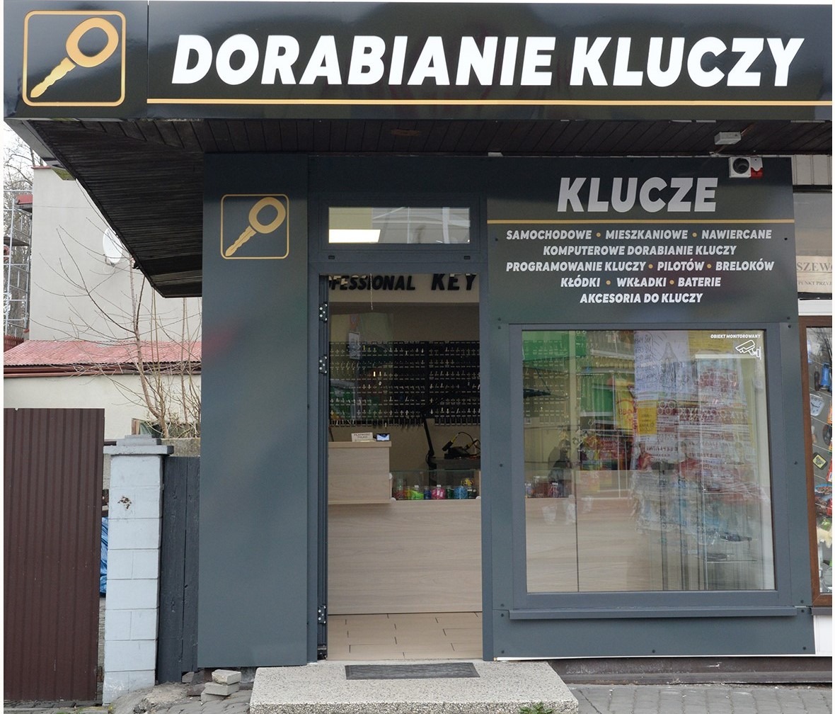 Fasada punktu dorabiania kluczy z szyldem 'Dorabianie Kluczy', widoczne liczne klucze wewnątrz oraz oferta usług: klucze samochodowe, mieszkaniowe, nawiercane, komputerowe dorabianie kluczy...