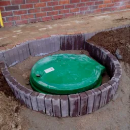 Zielona pokrywa studzienki otoczona półkolem z kostki brukowej w kolorze czerwono-szarym, widoczny fragment ceglanego muru i ziemia.