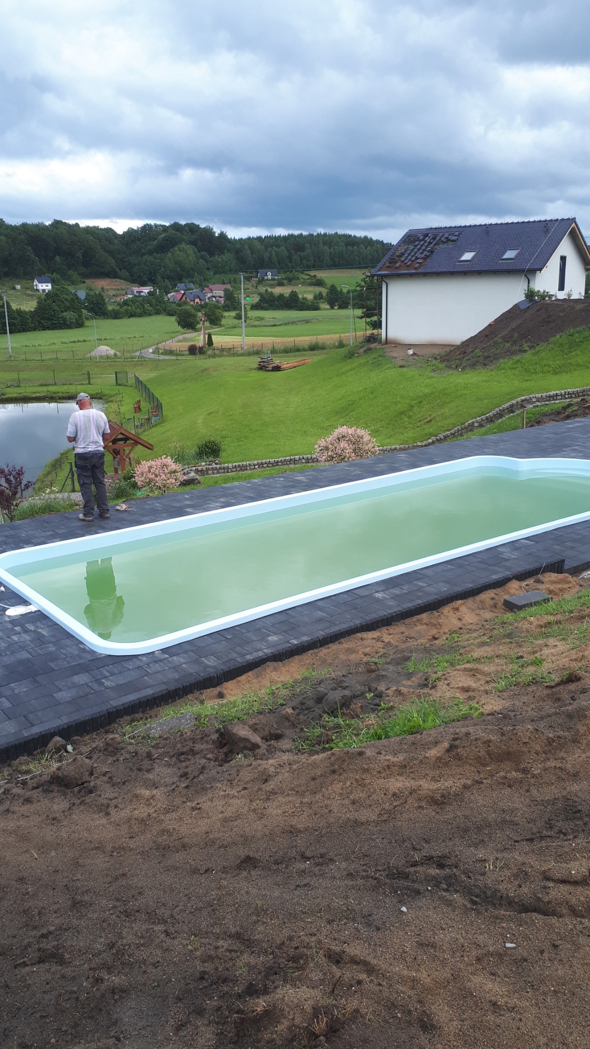 Niebieski basen z wodą otoczony ciemnoszarą kostką brukową w ogrodzie, widok na budynek z ciemnym dachem i zielone wzgórza w tle, mężczyzna przy basenie.