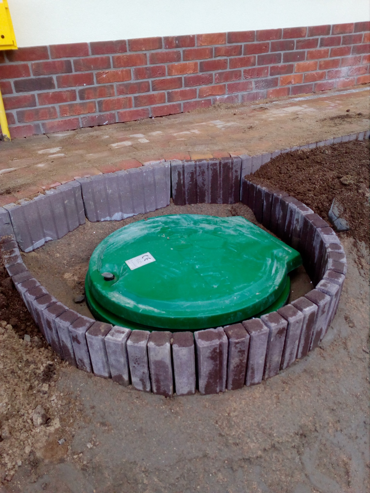 Zielona pokrywa studzienki otoczona półkolem z kostki brukowej w kolorze czerwono-szarym, widoczny fragment ceglanego muru i ziemia.