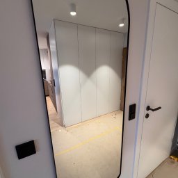 Ir Home Handicrafts Tomaszewski Andrzej - Wnętrze mieszkania z widoczną szafą wnękową w lustrze o nieregularnym kształcie. Obok białe drzwi z czarną klamką. Na ścianie widoczny włącznik światła.