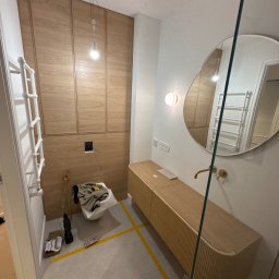 Ir Home Handicrafts Tomaszewski Andrzej - Nowoczesna łazienka w trakcie wykończenia: drewniana zabudowa ściany, szafka pod umywalkę z frezowanym frontem, okrągłe lustro. Widoczne narzędzia i taśmy malarskie.