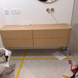 Ir Home Handicrafts Tomaszewski Andrzej - Montaż szafki łazienkowej z drewnianymi frontami, poziomowana laserowo. Widoczne akcesoria montażowe i plan. Umywalka z baterią w kolorze złotym.